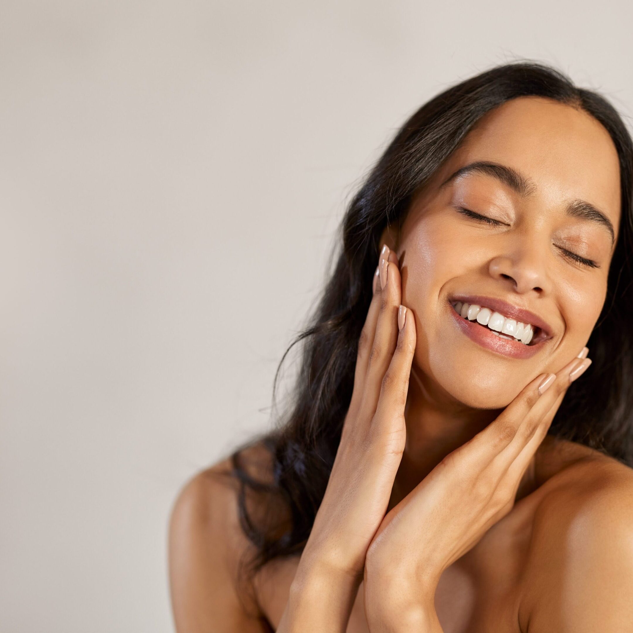 Portrait of beautiful latin woman touching her clean and healthy face against grey background. Smiling hispanic woman with natural makeup feeling healthy skin with eyes closed. Attractive multiethnic young woman pampering with perfect skin isolated with copy space.