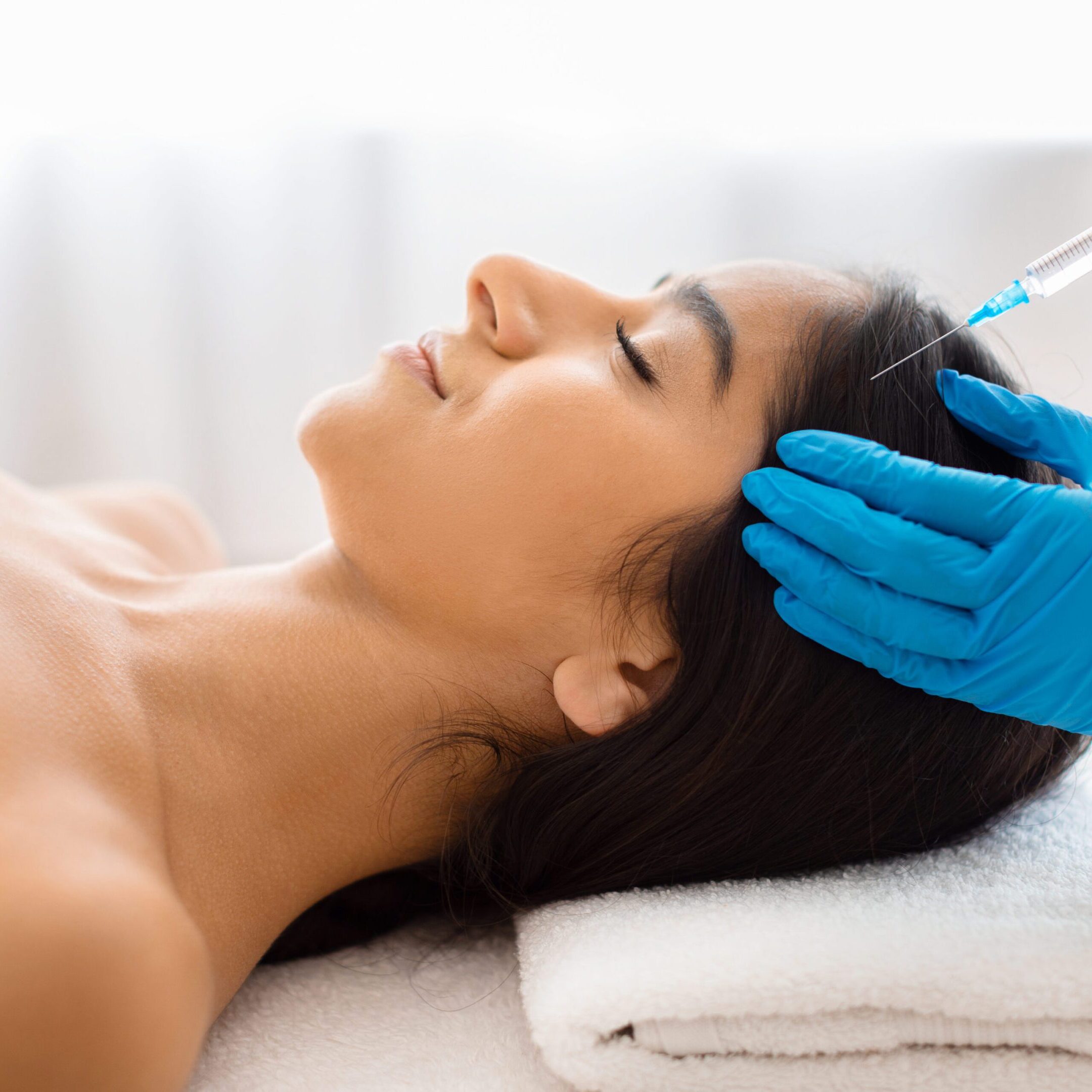Beautician making scalp injections for smiling young indian lady Hair loss, alopecia treatment. Unrecognizable beautician wearing medical gloves holding syringe, making scalp injections for smiling young indian lady, SPA beauty salon interior