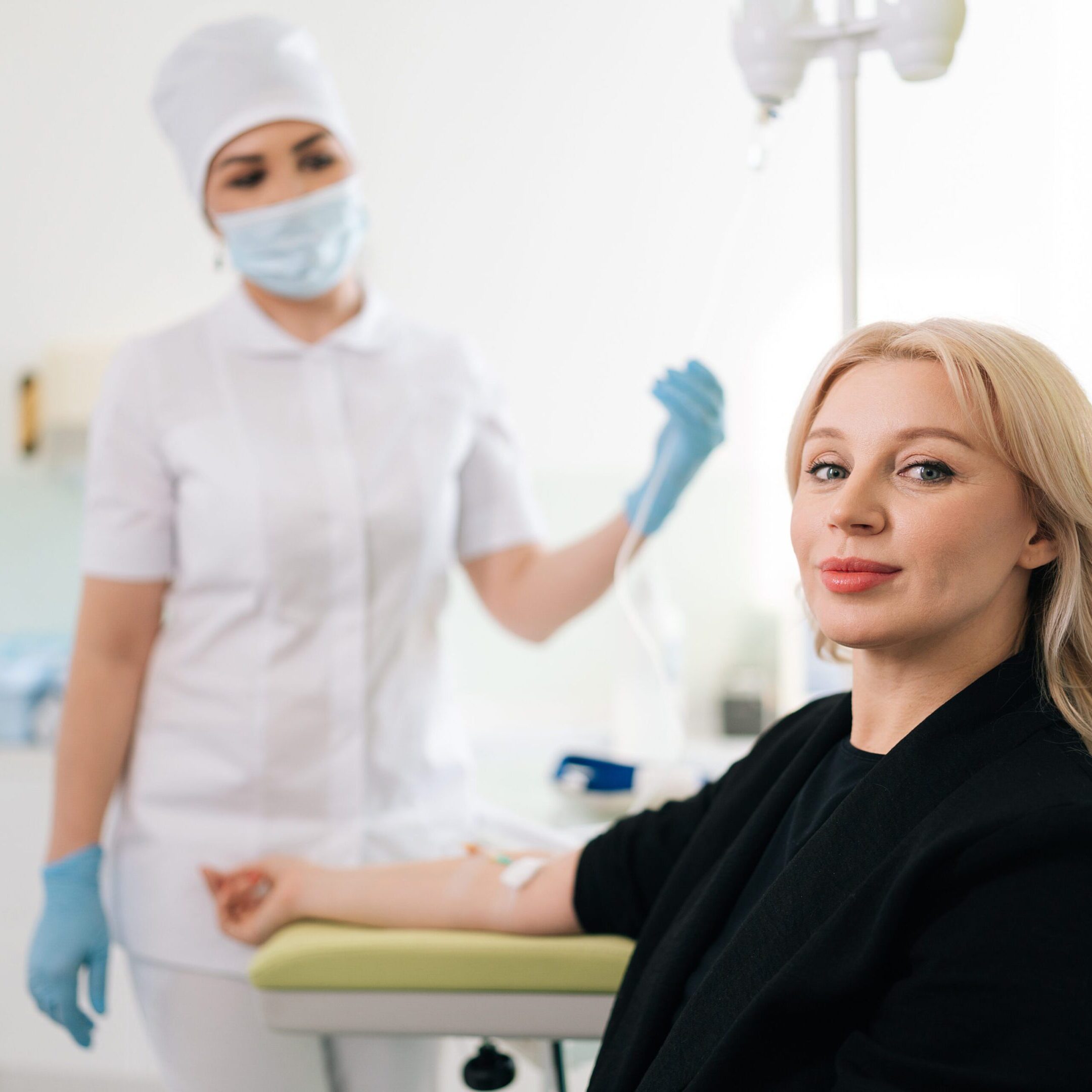 Portrait of blonde woman sitting on medical chair and getting intravenous vitamin therapy looking at camera. Clinic nurse in uniform standing on blurred background. Healthcare, wellness concept.
